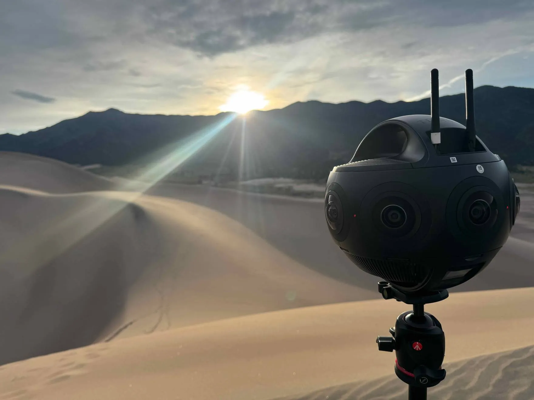 A 360-degree capture camera on a tripod overlooking sand dunes and mountains at golden hour