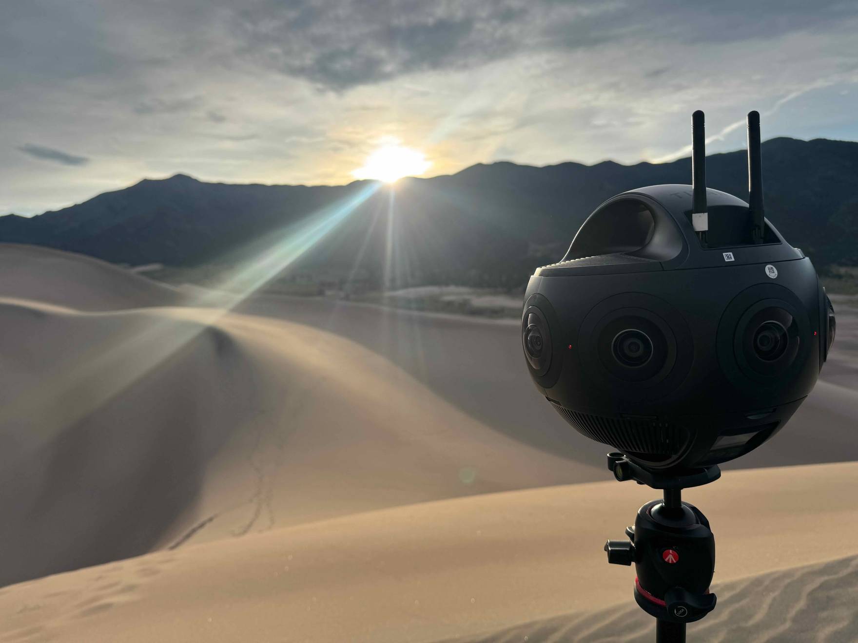 A 360-degree capture camera on a tripod overlooking sand dunes and mountains at golden hour